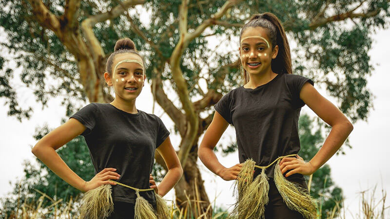 Two Indigenous youths in costume standing in front of a tree